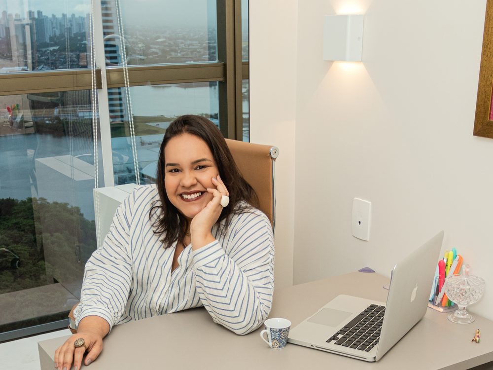Mayara sorrindo à mesa do consultório, com vista para Recife