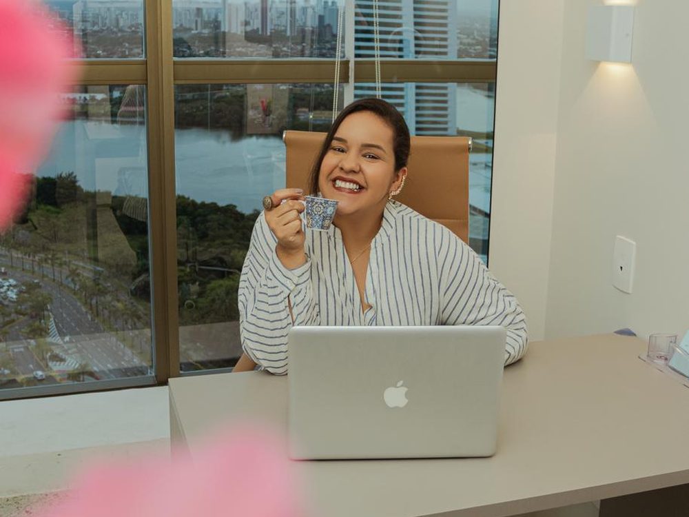 Mayara sorrindo com uma xícara de café, em frente ao notebook no consultório